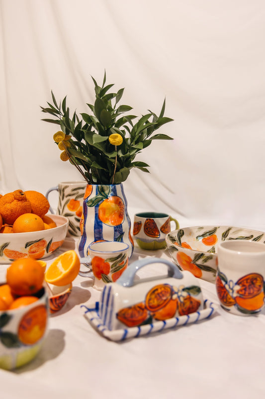 Hand-painted ceramic tableware featuring bright orange fruit designs, including bowls, mugs, a vase with greenery, and fresh oranges on a white backdrop.